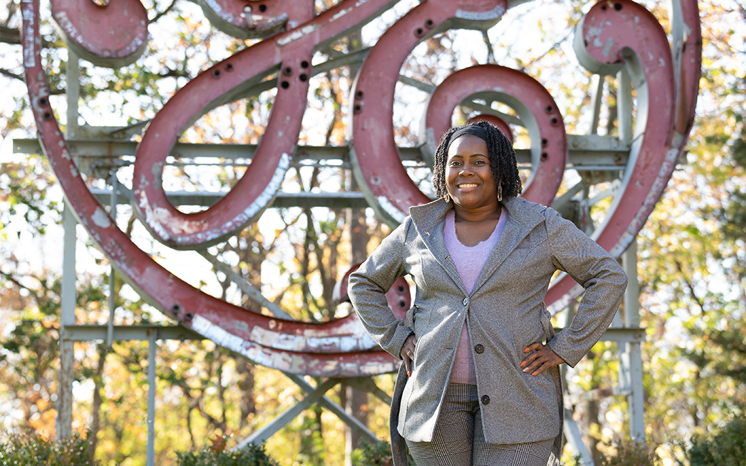 A person stands confidently with hands on hips in front of a large, vintage GE sign. They are wearing a gray blazer, pink shirt, and checkered pants. The background features autumn trees and greenery.